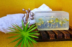 A decorative Dirty Soap 24K Gold Bar Soap with the word "bath" on a wooden rack, accompanied by towels, a green plant, and ornamental spheres against a 24K Gold background.