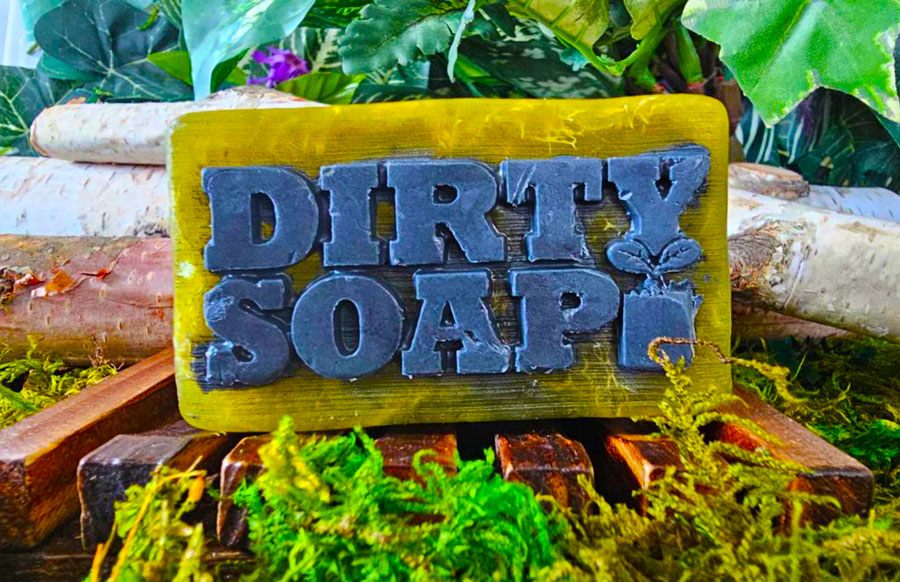 A colorful Gentlemen's Choice Bar Soap with the words "dirty soap" embossed on it, surrounded by a rustic natural setting with wood and moss.