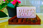 A novelty bar soap labeled "Girl's Undercover Bar Soap," crafted from a natural blend including Goat's Milk, placed on a wooden rack with a potted plant beside it.