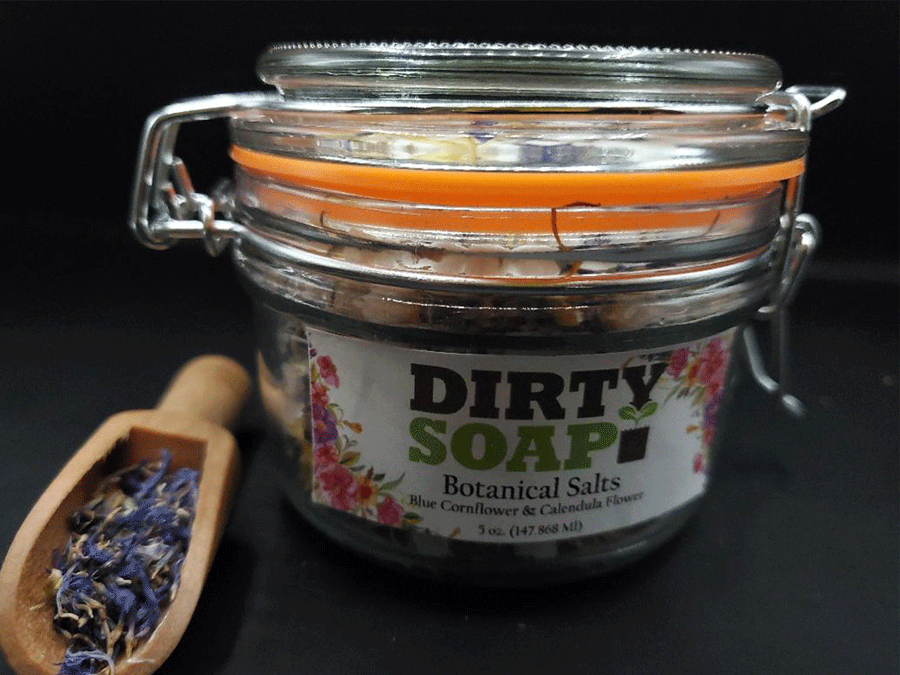 A glass jar with a label that reads "Dirty Soap Blue Cornflower and Calendula Flower" beside a wooden scoop with dried lavender.