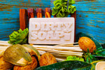 A bar of Eucalyptus and Spearmint Bar Soap labeled "Dirty Soap Bubbles" surrounded by natural ingredients and cinnamon sticks on a blue wooden background.