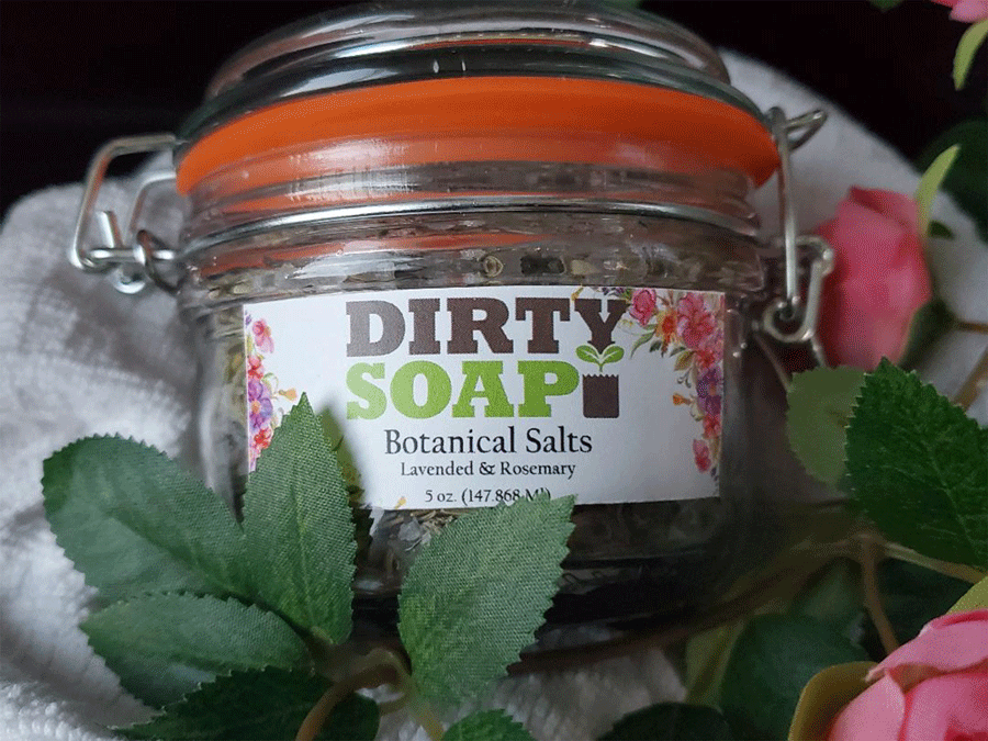 A jar of Dirty Soap Lavender Rosemary Botanical Salt, surrounded by green leaves and pink flowers.