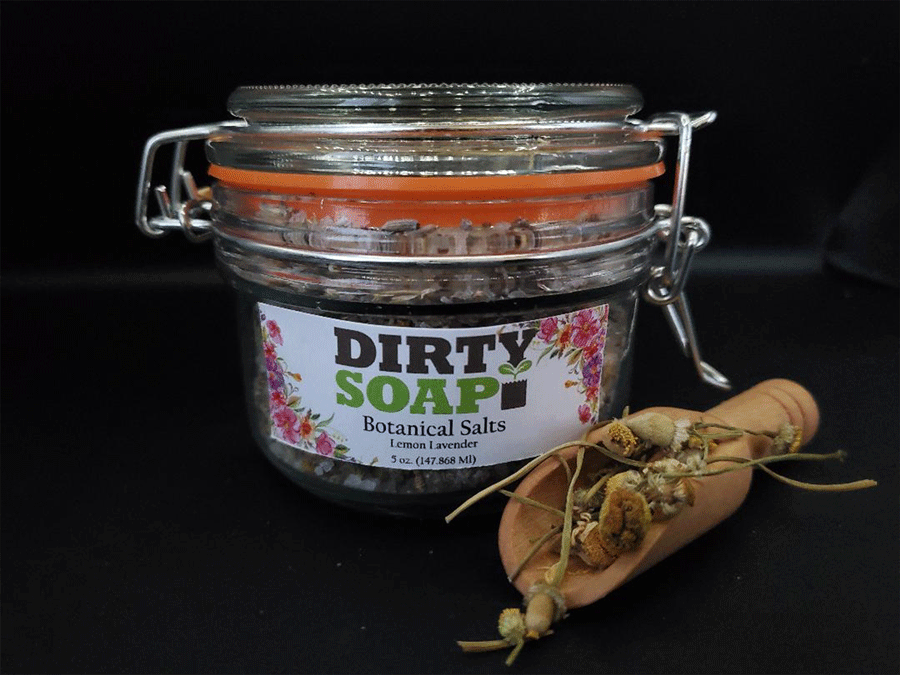 A jar of "Dirty Soap Lemon Lavender Botanical Salt" beside a small wooden scoop with dried botanicals.