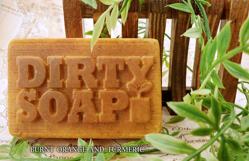 A bar of Burnt Orange & Turmeric with Safflower Oil handmade soap from Dirty Soap Bubbles, with the words "dirty soap" embossed on it, positioned against an earthy background with green leaves.