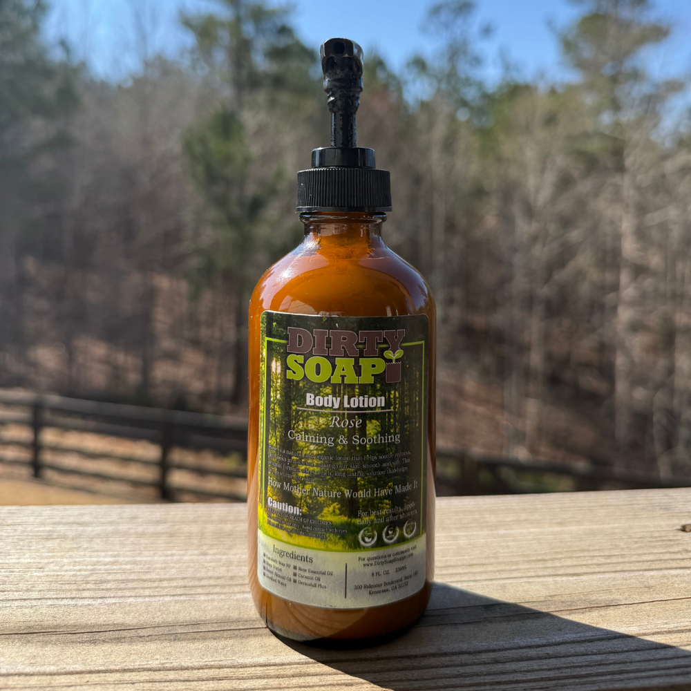 A bottle of Dirty Soap body lotion, rose scent, is placed on a wooden surface outdoors with trees and a fence in the background.