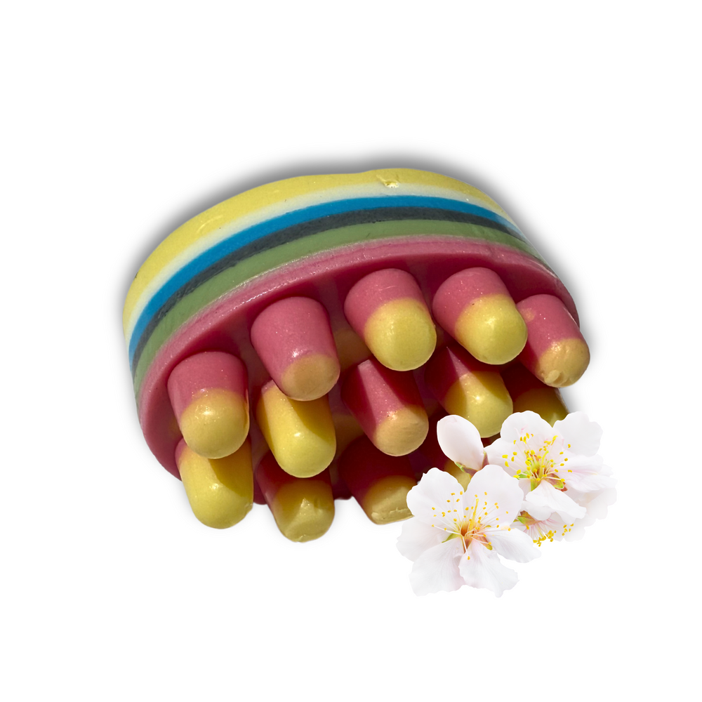 The Dirty Soap Blossom Massage Bar, infused with essential oils and featuring vibrant stripes with rounded pink and yellow accents, is artistically displayed next to white cherry blossom flowers on a white background.