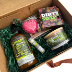 The Dirty Soap Gift Box Set by Dirty Soap includes body lotion, lip balm, soap bar, flower soap, and a heart-shaped soap—complete body care essentials elegantly arranged on green shredded paper.