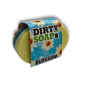 The Blossom Massage Bar by Dirty Soap is a yellow oval soap with a blue wrapper decorated with white daisies and green text, photographed on a white background—infused with essential oils for relaxation.
