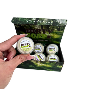 A hand holds a tin labeled "Dirty Soap" above an open Whipped Body Butter Sampler by Dirty Soap, featuring four unique body butter scents—ideal for anyone looking to elevate their skincare routine.