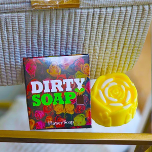 A box labeled "Dirty Soap Shoppe" with a floral design sits next to a handmade Yellow Rose Honeysuckle & Jasmine Flower Soap, shaped like a rose, on a glass table.