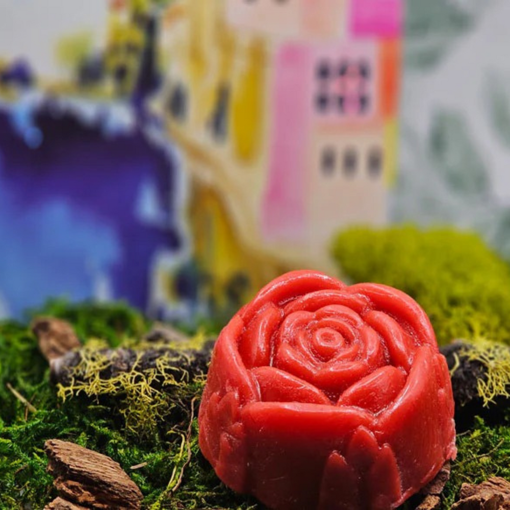 The Dirty Soap Shoppe's Red Rose Flower Soap, handmade and rose-shaped, is infused with essential oils and displayed on green moss and wood, set against a vibrant, abstract backdrop.