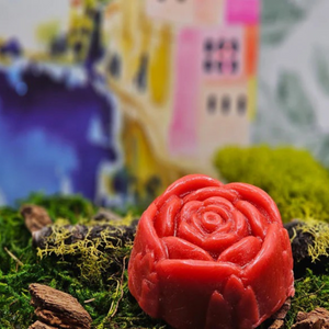 The Dirty Soap Shoppe's Red Rose Flower Soap, handmade and rose-shaped, is infused with essential oils and displayed on green moss and wood, set against a vibrant, abstract backdrop.