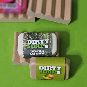 Two Dirty Soap bars—one eucalyptus & spearmint, the other mango & sand—sit on eco-friendly Mini Soap Dishes by Dirty Soap, made from natural pine wood and displayed on a green surface.