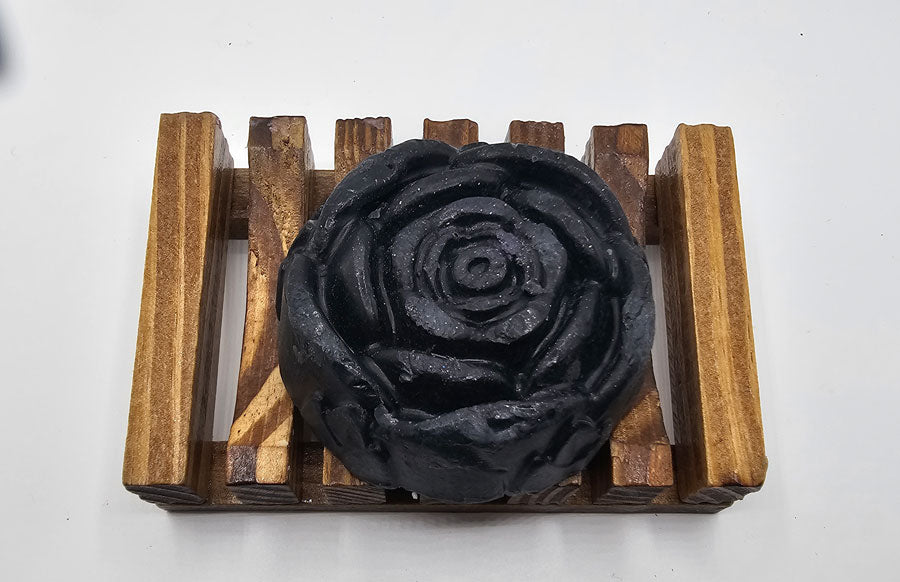 A handmade Dirty Soap Shoppe Black Rose Peppermint Flower Soap, elegantly shaped like a rose, is placed on a rectangular wooden soap dish with horizontal slats, set against a pristine white background. This unique soap offers not just aesthetic beauty but also a soothing effect.