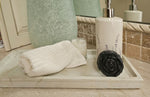 A bathroom countertop with a white towel, a small lit candle, a handmade Black Rose Peppermint Flower Soap from Dirty Soap Shoppe, and a white soap dispenser on a tray.