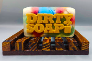 A colorful handmade Christmas Rocks bar of soap from Dirty Soap, with the words "dirty soap" embossed on it, resting on a wooden soap dish.