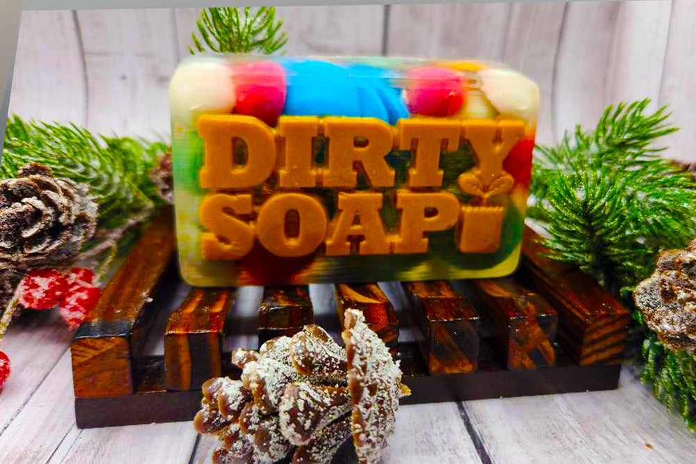 Multicolored handmade Christmas Rocks bar of soap with the words "Dirty Soap" embossed on it, displayed on a wooden soap dish with pine cones and greenery around.