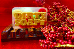 A bar of soap labeled "Christmas Rocks Traditional Bar Soap" infused with Lavender Essential Oil, presented on a wooden holder against a red background with decorative red berries. Brand name: Dirty Soap