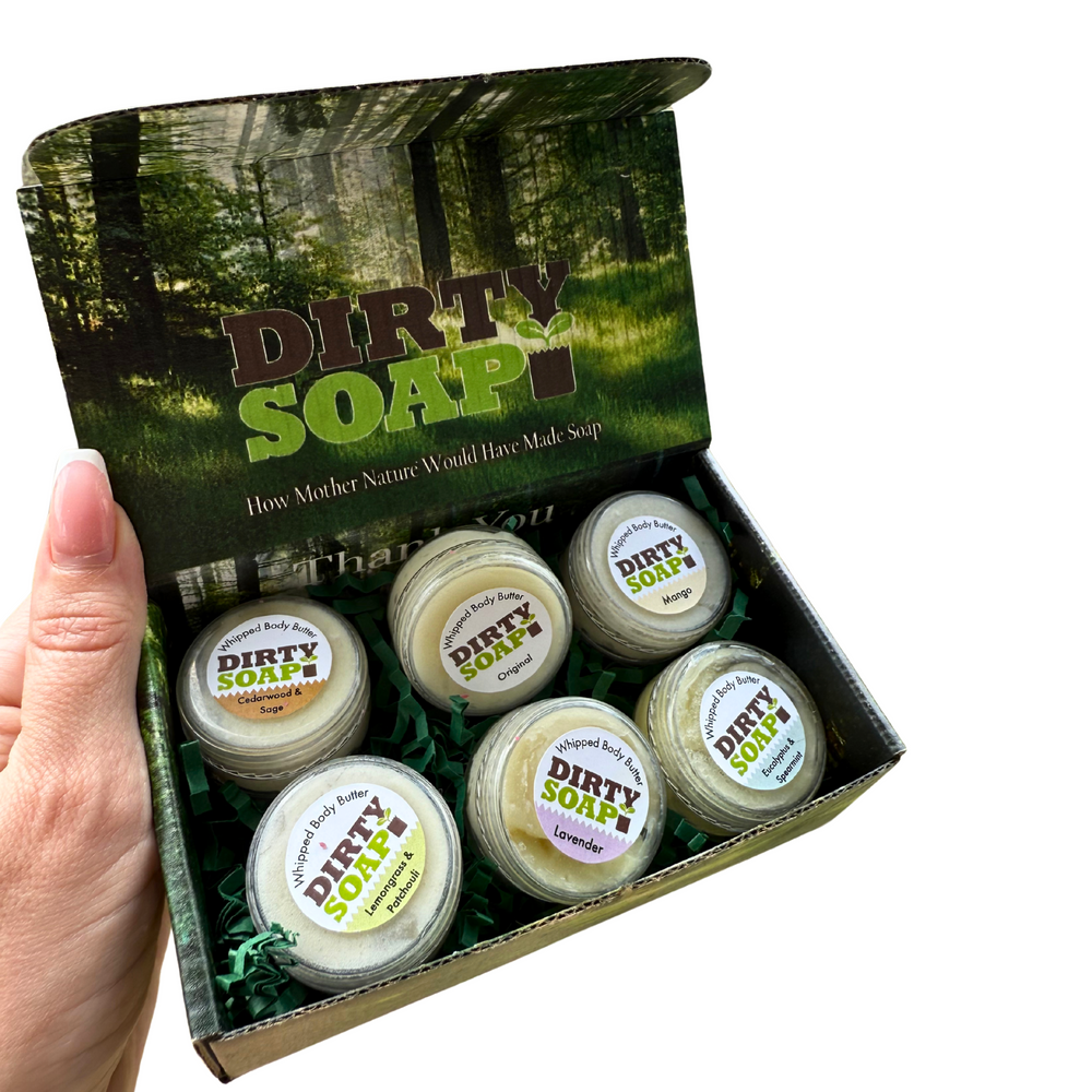 A hand holds an open Dirty Soap Whipped Body Butter Sampler box, showing six tubs of body butter in assorted scents against a forest-inspired background.