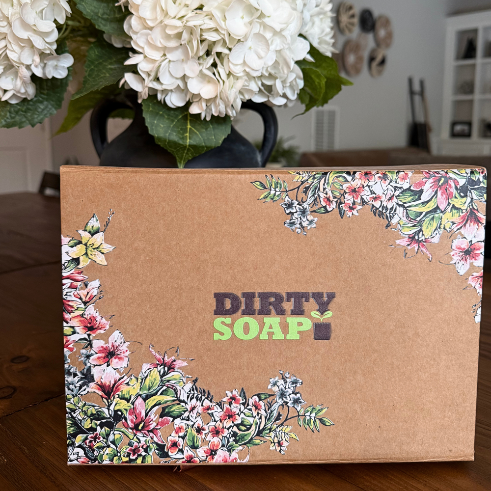 The Dirty Soap Gift Box Set by Dirty Soap, featuring floral designs, rests on a wooden table surrounded by white flowers—a charming addition to your body care essentials.