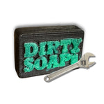 A black bar of The Mechanic Bar Soap by Dirty Soap Bubbles, labeled "DIRTY SOAP" in green, with exfoliating pumice, rests beside a silver adjustable wrench on a white background.