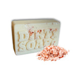A beige rectangular Sunrise Grapefruit & Coconut with Himalayan Salt Bar Soap by Dirty Soap Bubbles, featuring embedded pieces of pink Himalayan salt, is shown next to a small pile of loose salt crystals.