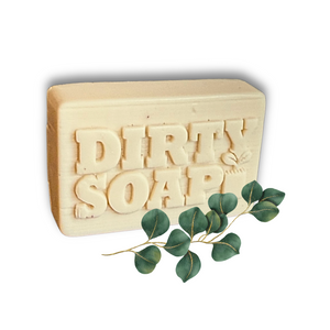 A beige Eucalyptus and Spearmint Bar Soap from Dirty Soap Bubbles, embossed with "DIRTY SOAP," sits beside eucalyptus and spearmint leaves on a white background.