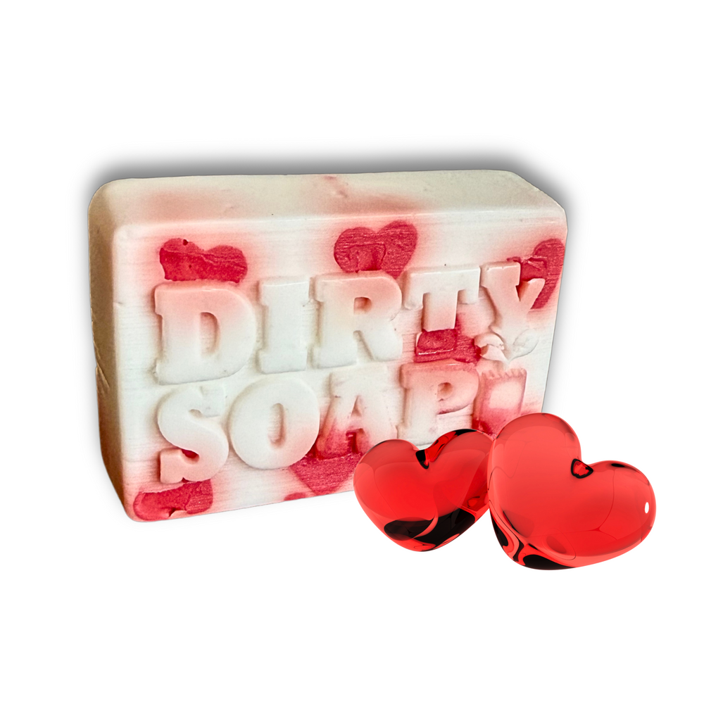 A rectangular Self-Care Bar Soap by Dirty Soap Bubbles, embossed with “DIRTY SOAP,” made with Goat’s Milk and decorated with red hearts, sits beside two glossy red heart-shaped objects.