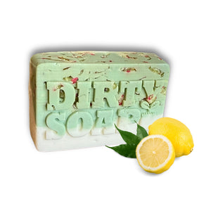 A green and white Lemon Squeeze Bar Soap from Dirty Soap Bubbles rests beside two whole lemons and a lemon half on a white background.