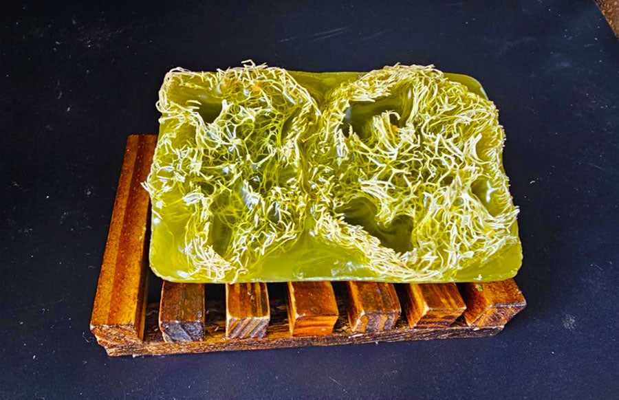 A translucent green bar of Gentlemen's Choice Bar Soap with a loofah embedded inside, displayed on a wooden holder by Dirty Soap.