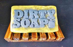 A handmade Gentlemen's Choice bar of soap engraved with the words "Dirty Soap" resting on a wooden soap dish.