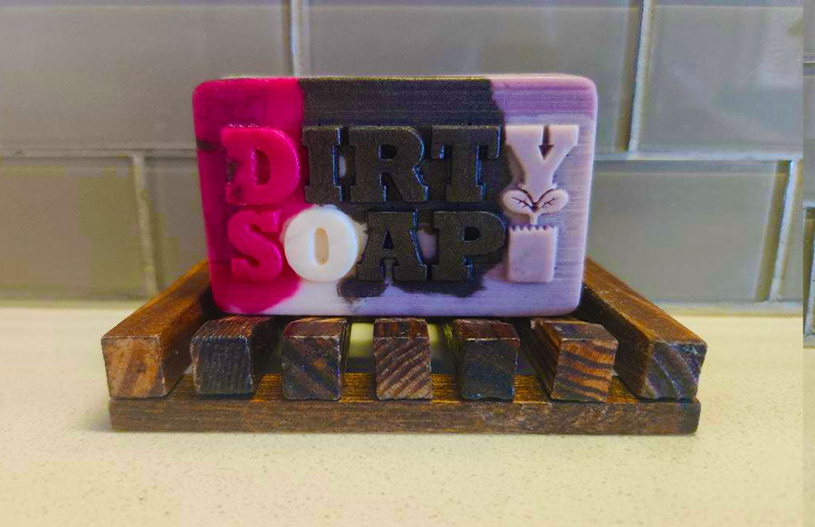 A multicolored bar of soap labeled "Girl's Undercover Bar Soap" by Dirty Soap, enriched with goat's milk and beet root powder, rests on a wooden soap dish on a countertop.