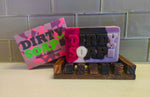 The "Girl's Undercover Bar Soap" by Dirty Soap features a handmade bar with "DIRTY SOAP" text and a rabbit design, placed on a wooden holder. The soap, made from Goat's Milk and essential oils, is showcased beside its vibrant camouflage-themed packaging labeled "DIRTY SOAP Girl Undercover," all against an elegant tiled background.