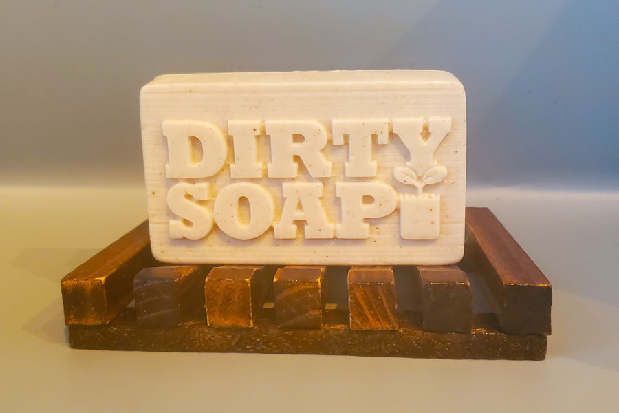 A Mango and Bergamot Bar Soap with the brand "Dirty Soap" embossed on it, resting on a wooden soap dish.