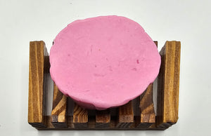 A round pink Pink Rose Jasmine Flower Soap bar from Dirty Soap Shoppe sits on a wooden soap dish against a white background. This handmade beauty not only enhances your bathing experience but also reduces dark spots for a clearer complexion.