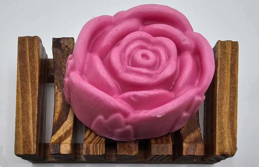 A handmade Pink Rose Jasmine Flower Soap from Dirty Soap Shoppe, shaped like a rose and placed on a wooden soap holder, not only adds elegance but also reduces dark spots.