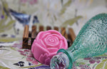 A pink, rose-shaped handmade soap sits next to a textured green glass bottle on a floral-patterned surface. A wooden soap dish is in the background. This Pink Rose Jasmine Flower Soap from Dirty Soap Shoppe not only enhances the aesthetic but also reduces dark spots for a flawless complexion.