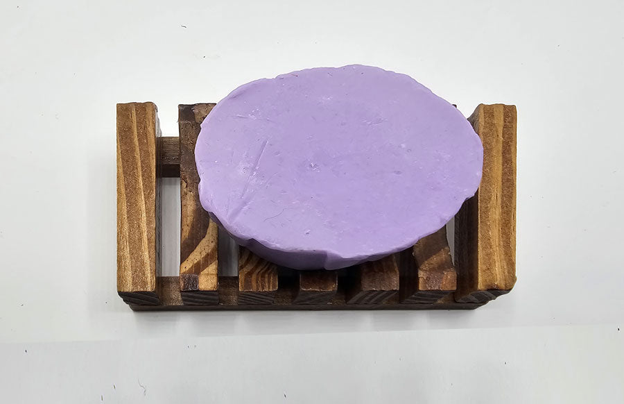 A round, Purple Rose Flower Soap by Dirty Soap Shoppe sits gracefully on a small wooden rack against a white background, showcasing its natural anti-inflammatory properties.