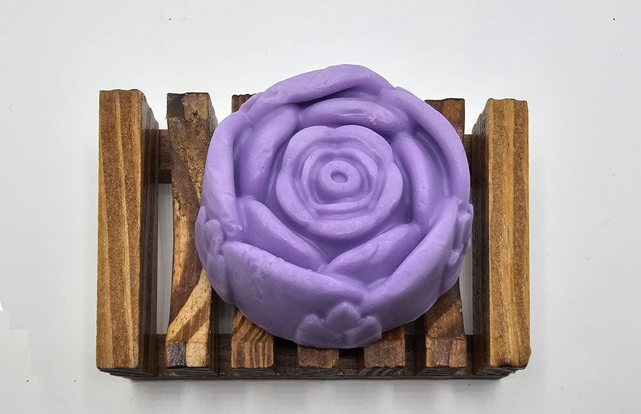 A Dirty Soap Shoppe Purple Rose Flower Soap, carefully handmade and shaped like a delicate blossom, rests on a wooden soap dish with parallel grooves against a white background. This artisan creation not only enchants with its beauty but also promises natural anti-inflammatory benefits for your skin.