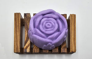 A Dirty Soap Shoppe Purple Rose Flower Soap, carefully handmade and shaped like a delicate blossom, rests on a wooden soap dish with parallel grooves against a white background. This artisan creation not only enchants with its beauty but also promises natural anti-inflammatory benefits for your skin.