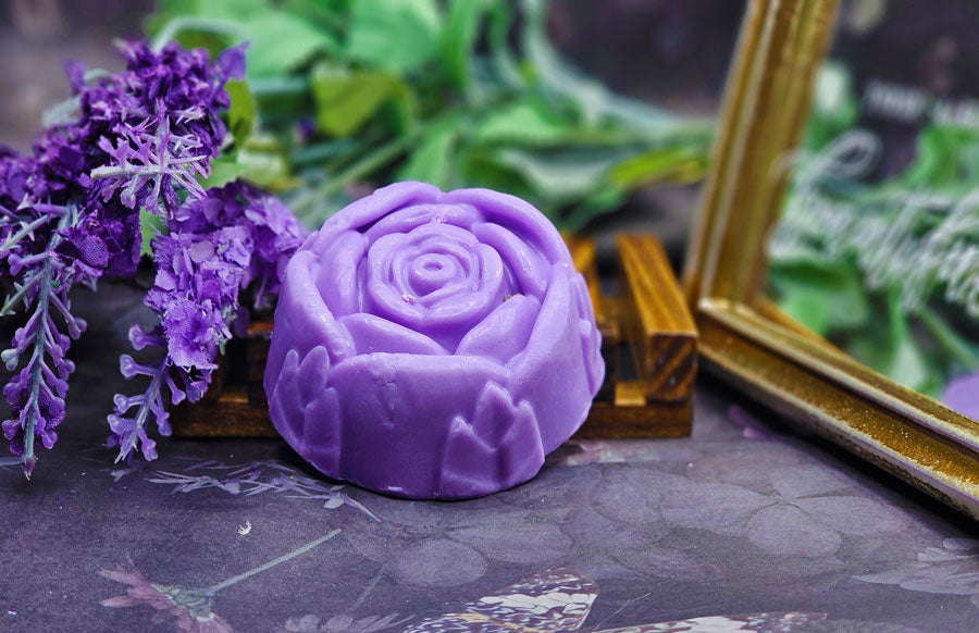 A Dirty Soap Shoppe Purple Rose Flower Soap shaped like a rose is displayed with lavender flowers and greenery in the background. A gold-framed sign is partially visible on the right, hinting at its soothing skin benefits and natural ingredients.