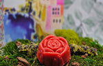 A Red Rose Flower Soap by Dirty Soap Shoppe, infused with spearmint essential oil, sits on green moss with a blurred, colorful background.