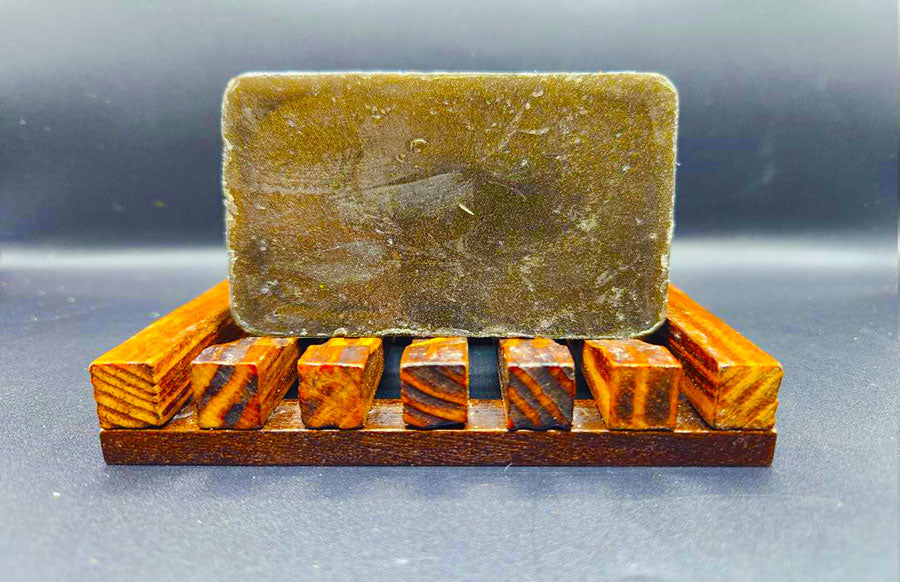 A bar of brown, handmade soap resting on a wooden soap dish. → The Mechanic Bar Soap by Dirty Soap Bubbles