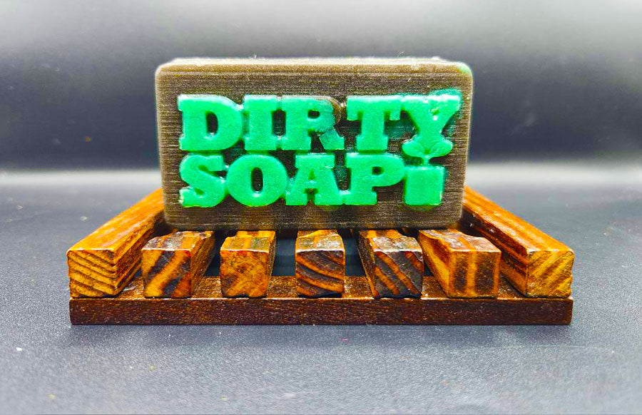 A handmade bar of The Mechanic Bar Soap with the words "dirty soap" embossed on it, resting on a wooden soap dish.