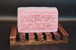 A bar of Dirty Soap's The New Pink Bar Soap with the words "dirty soap" embossed on it, resting on a wooden soap dish.