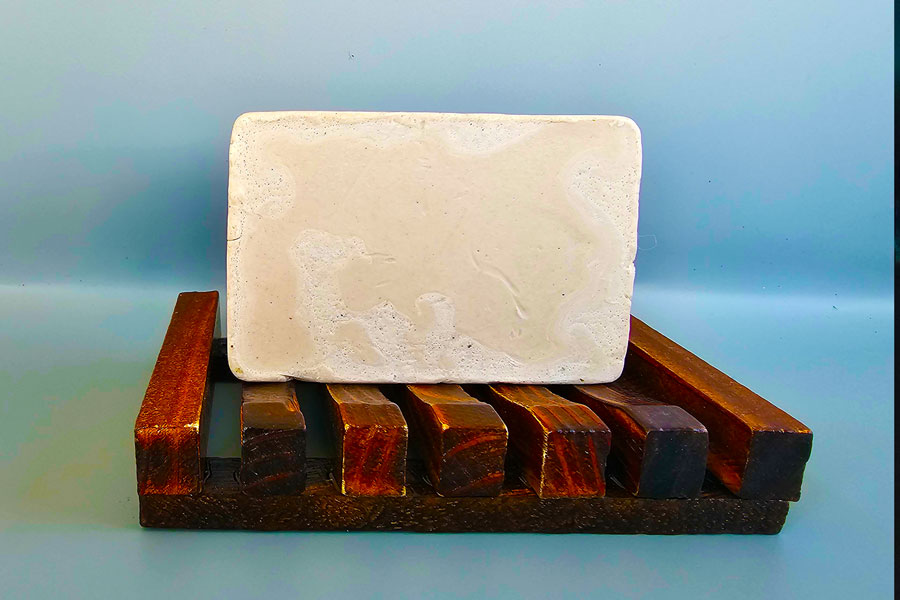 A Vanilla And Lavender Bar Soap from Dirty Soap resting on a wooden soap dish against a light blue background.