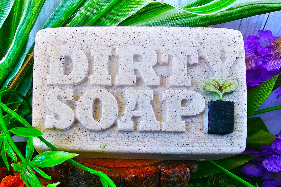 A bar of Dirty Soap's Vanilla And Lavender Bar Soap with the words "dirty soap" embossed on it, surrounded by greenery and purple flowers.