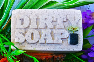 A bar of Dirty Soap's Vanilla And Lavender Bar Soap with the words "dirty soap" embossed on it, surrounded by greenery and purple flowers.