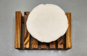 A round white cheese resting on a wooden rack against a gray background, reminiscent of the pure and calming aesthetics of Dirty Soap Shoppe’s White Rose Gardenia Flower Soap.