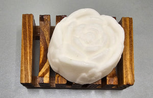 A White Rose Gardenia Flower Soap by Dirty Soap Shoppe on a wooden soap dish with a slatted design against a light grey background.